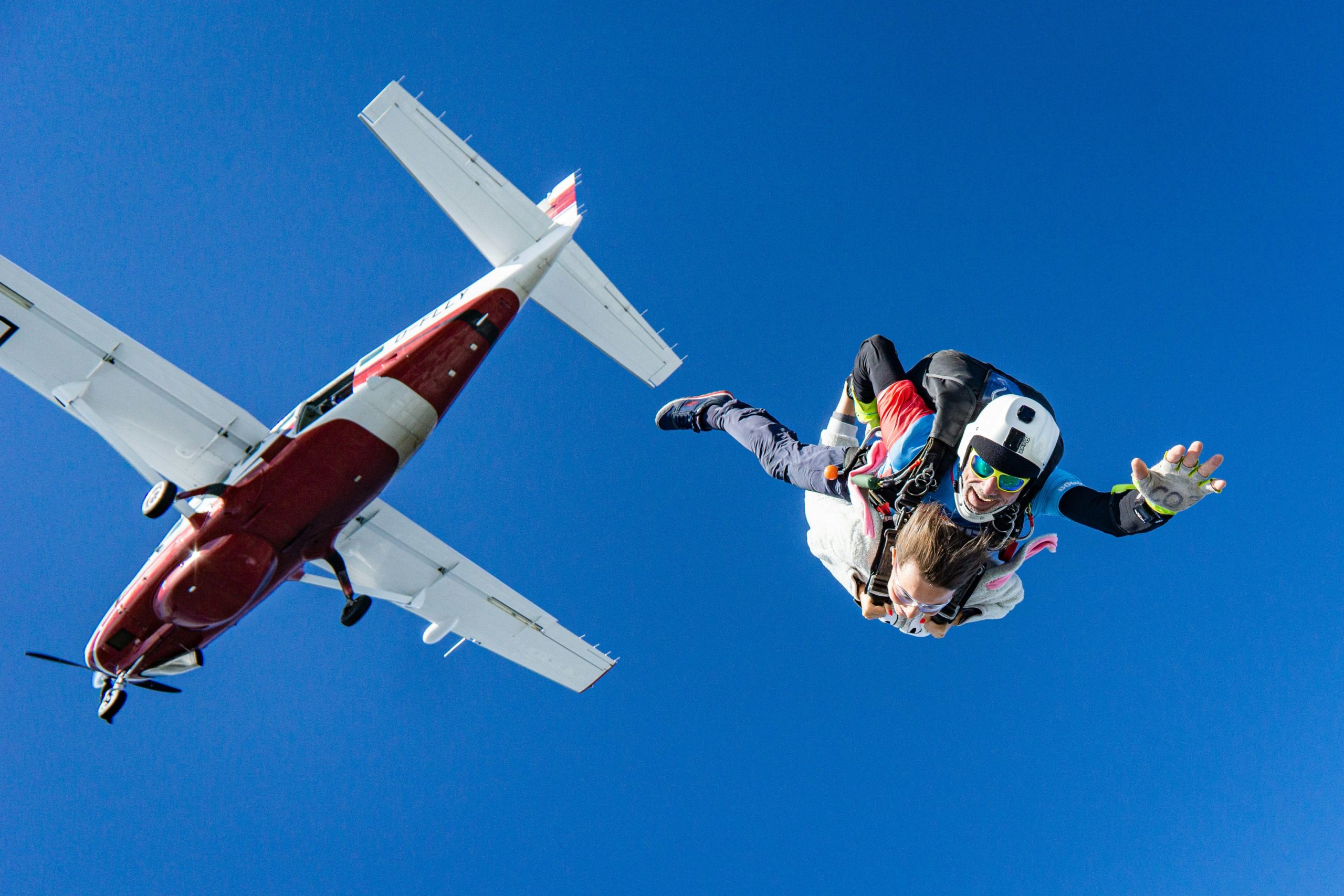Un parachutiste en combinaison de saut multicolore et casque blanc sourit à la caméra en tombant librement avec un petit avion rouge et blanc avec les ailes au-dessus de lui sur un ciel bleu clair.