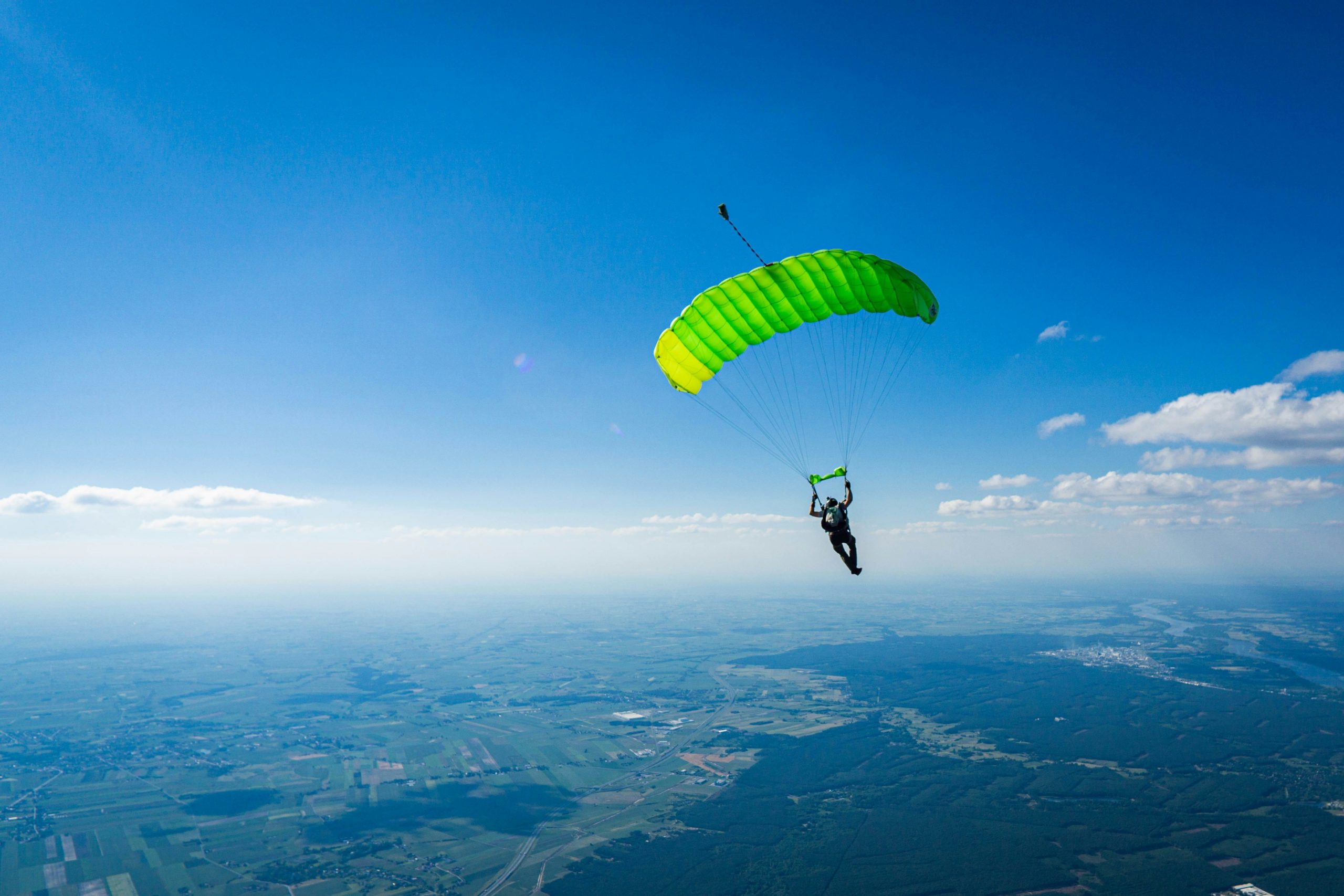 Parachutiste en descente avec un parachute vert fluorescent sous un ciel bleu clair au-dessus du paysage terrestre.
