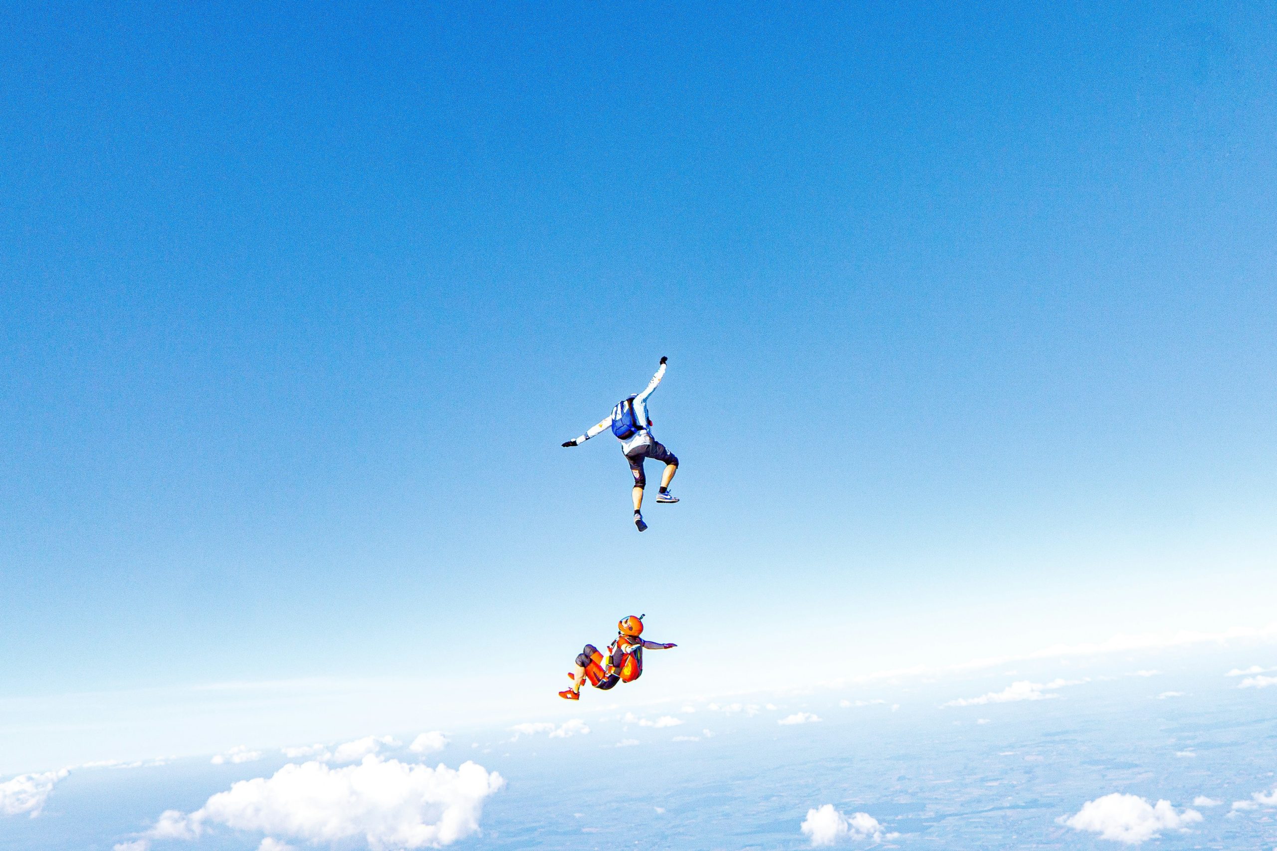 Un parachutiste en chute libre avec un bras levé et l'autre tendu vers le bas, et un second parachutiste en position horizontale en dessous, le tout sur un fond de ciel bleu clair parsemé de nuages et de la terre en vue lointaine en dessous.