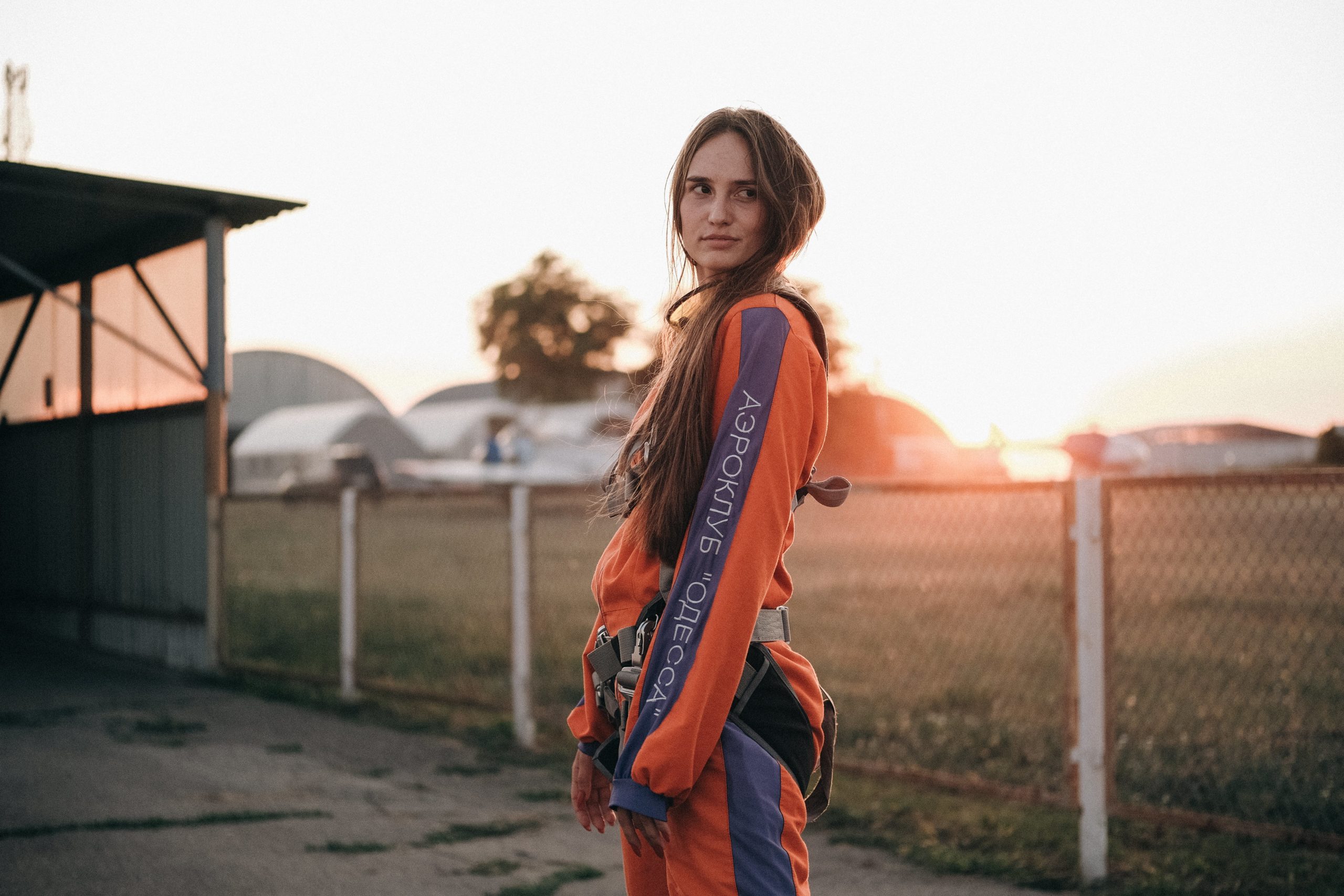  Une jeune femme en combinaison de parachutisme regarde par-dessus son épaule, devant un coucher de soleil sur un aérodrome.