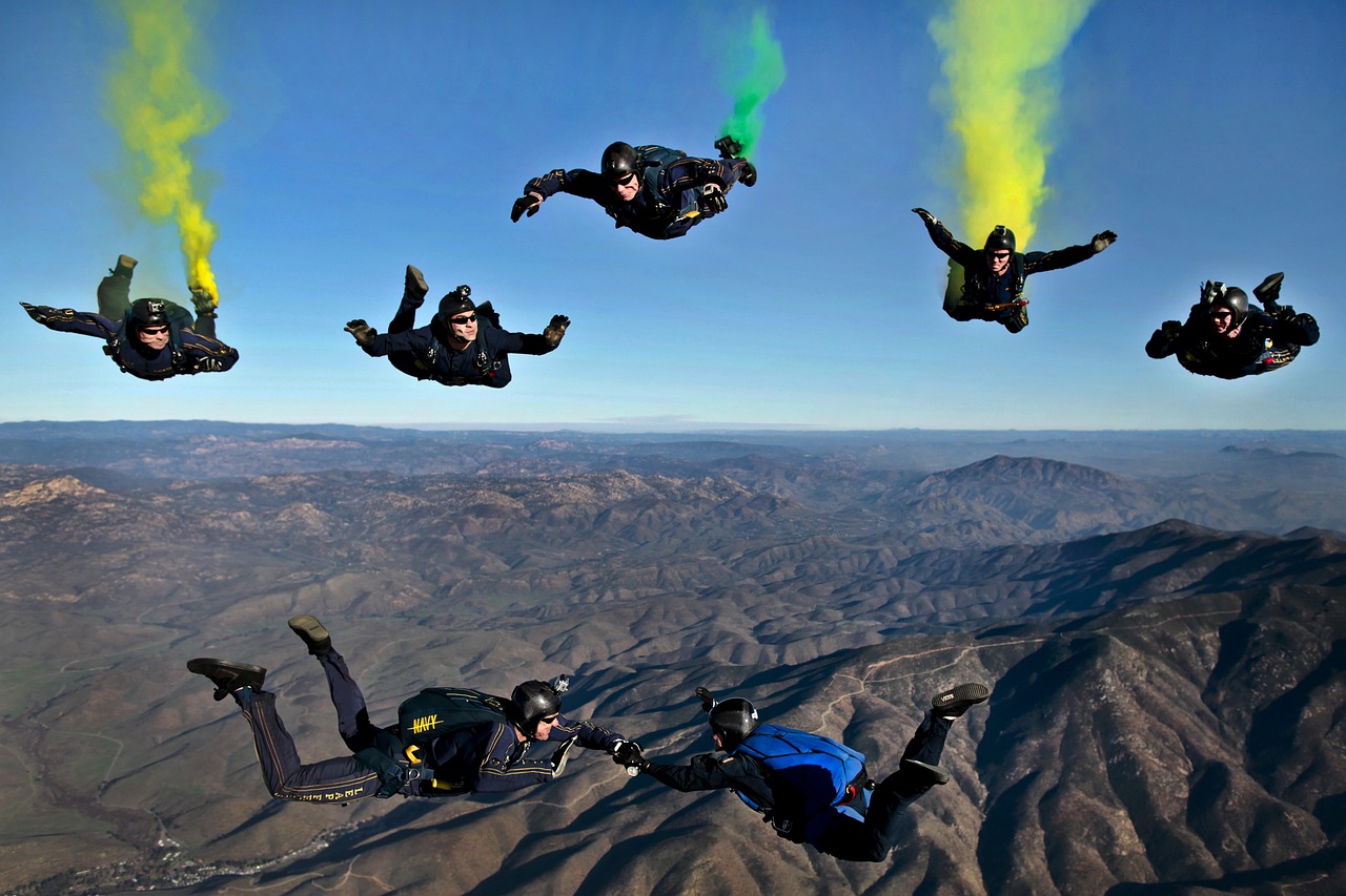 Six parachutistes en chute libre en formation, avec des fumigènes de couleur jaune et verte derrière eux, au-dessus d'un paysage montagneux