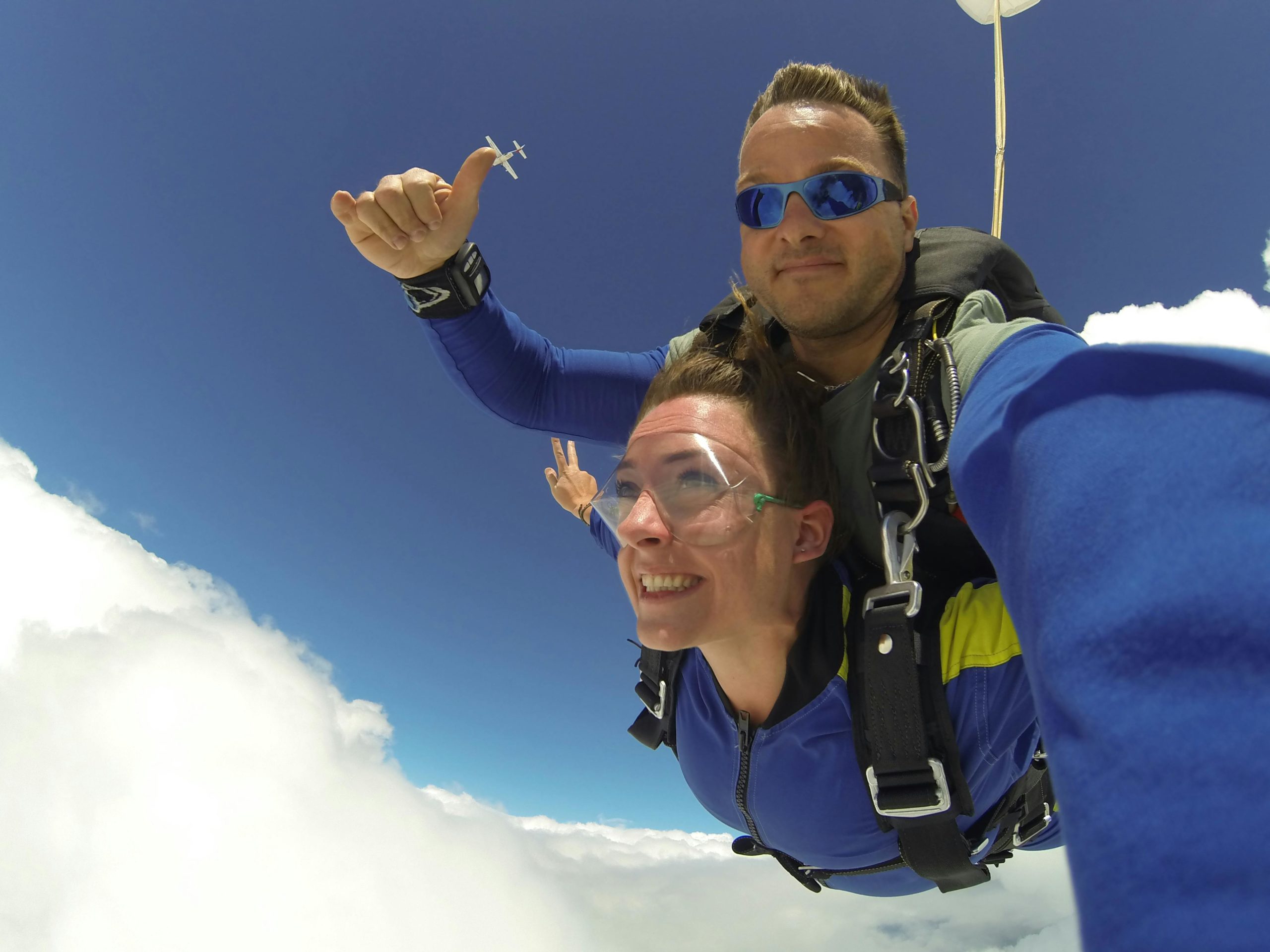 Autoportrait en chute libre de deux personnes faisant un saut en tandem en parachute, avec un instructeur derrière et un passager devant faisant le signe de la victoire, au-dessus des nuages avec un ciel bleu en arrière-plan.