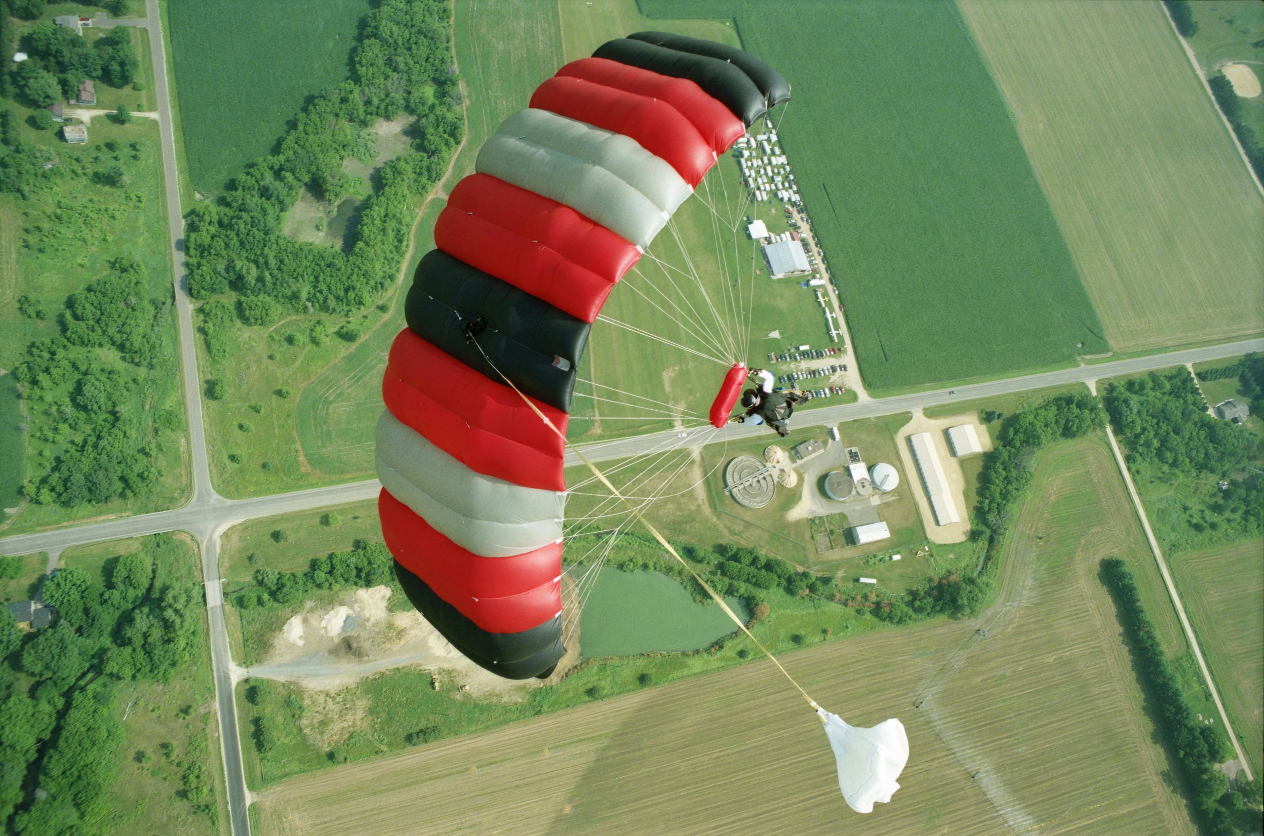 Une vue plongeante sur un parachutiste en plein vol avec un parachute rouge et noir, au-dessus d'un paysage verdoyant